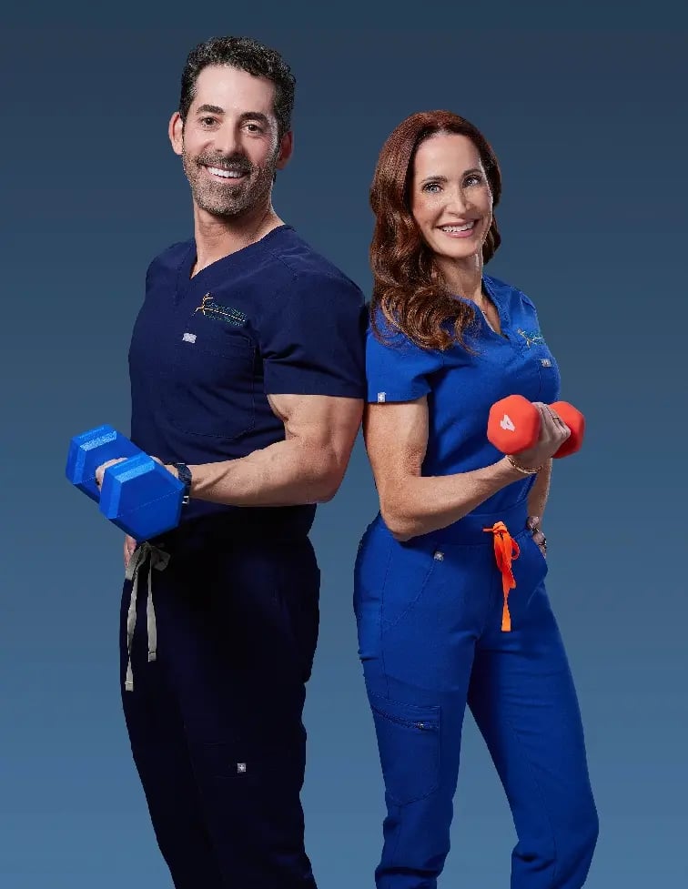 Dr Brad and Susan Sorosky in scrubs smiling, each holding dumbbells, promoting wellness and strength in a clinical setting.