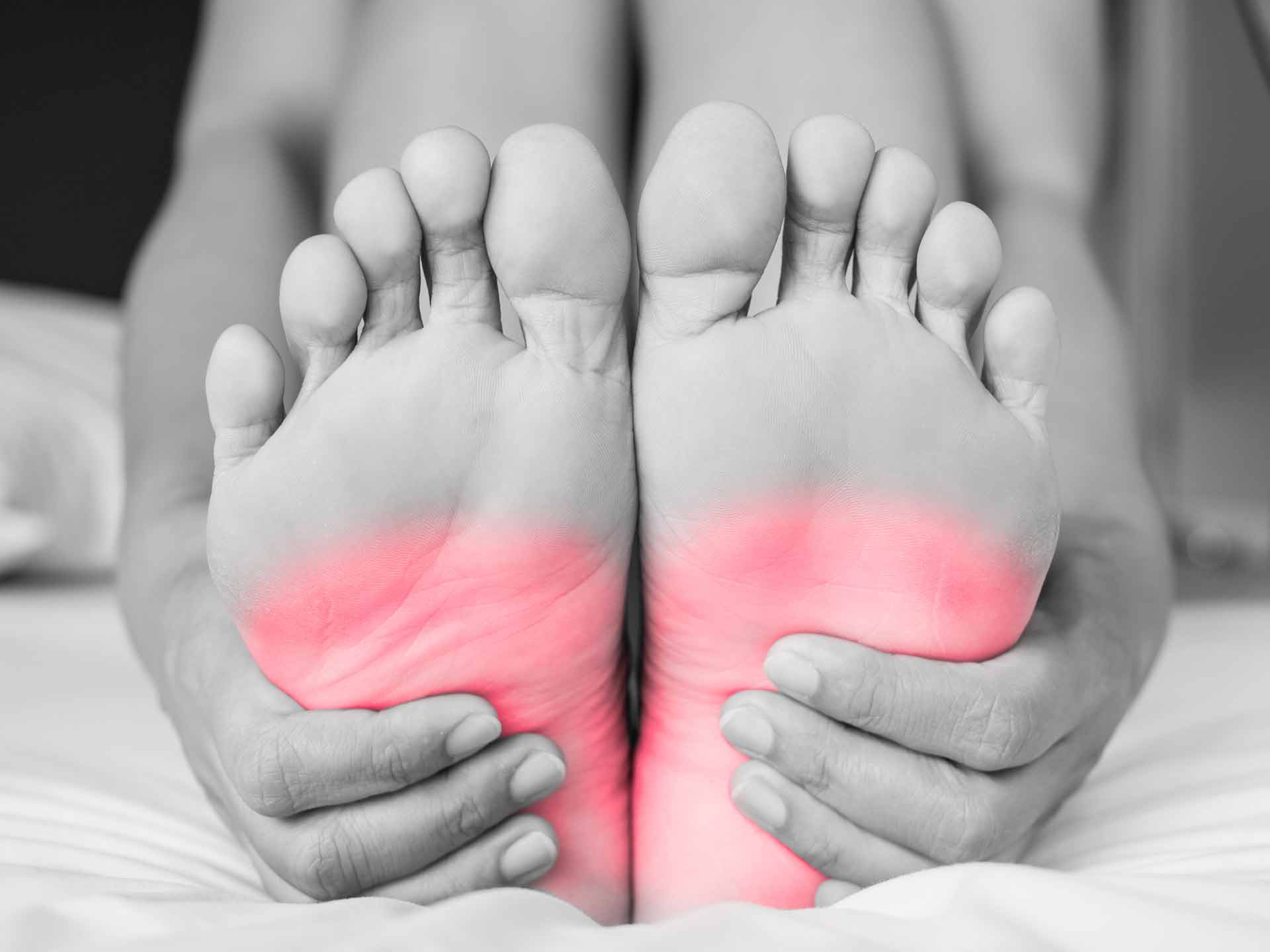 Black and white image of a person holding their feet with red highlighted areas on the soles, suggesting pain or discomfort